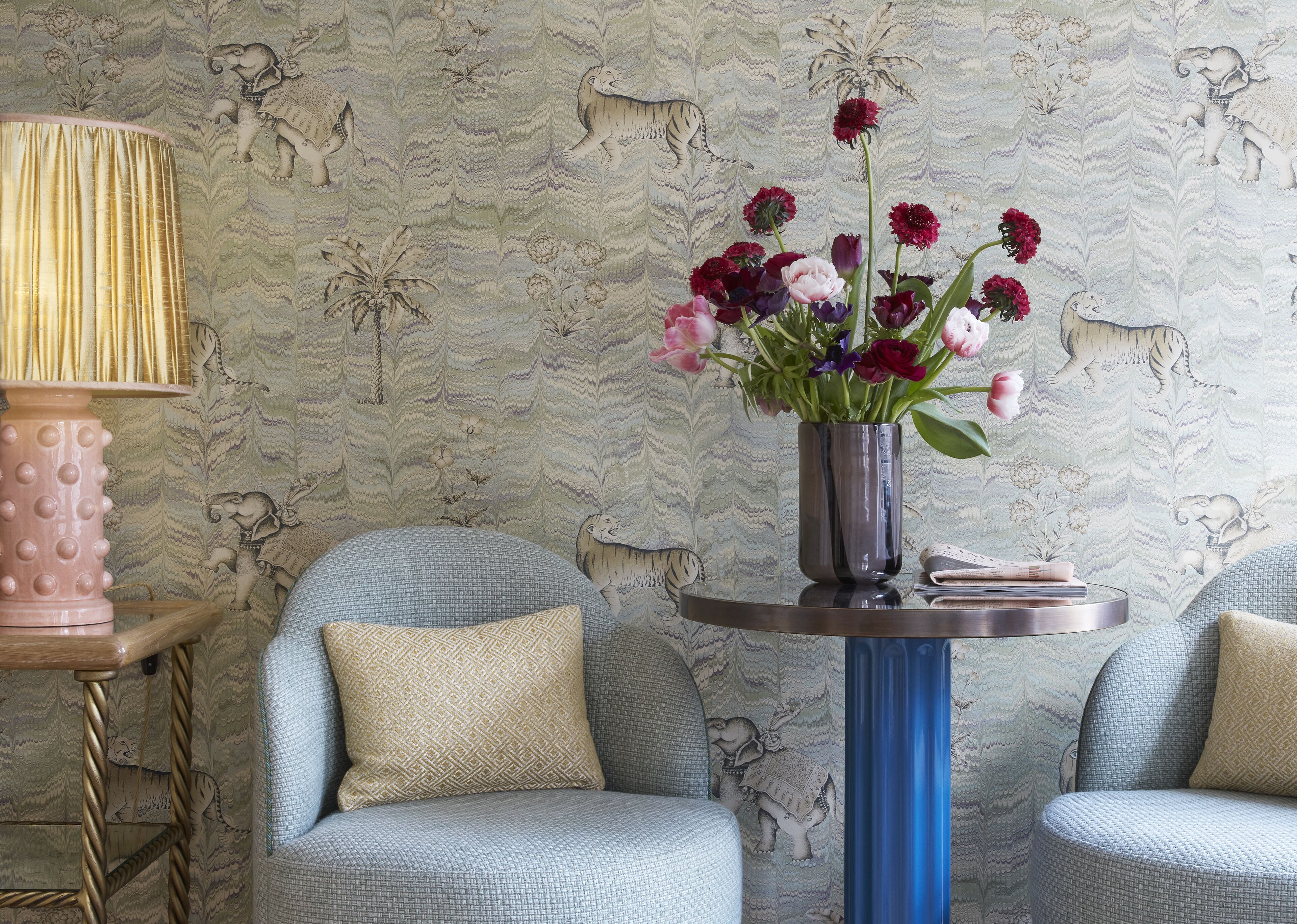 Um quarto com papel de parede estampado, uma mesa redonda com um vaso de flores, duas poltronas e um abajur.