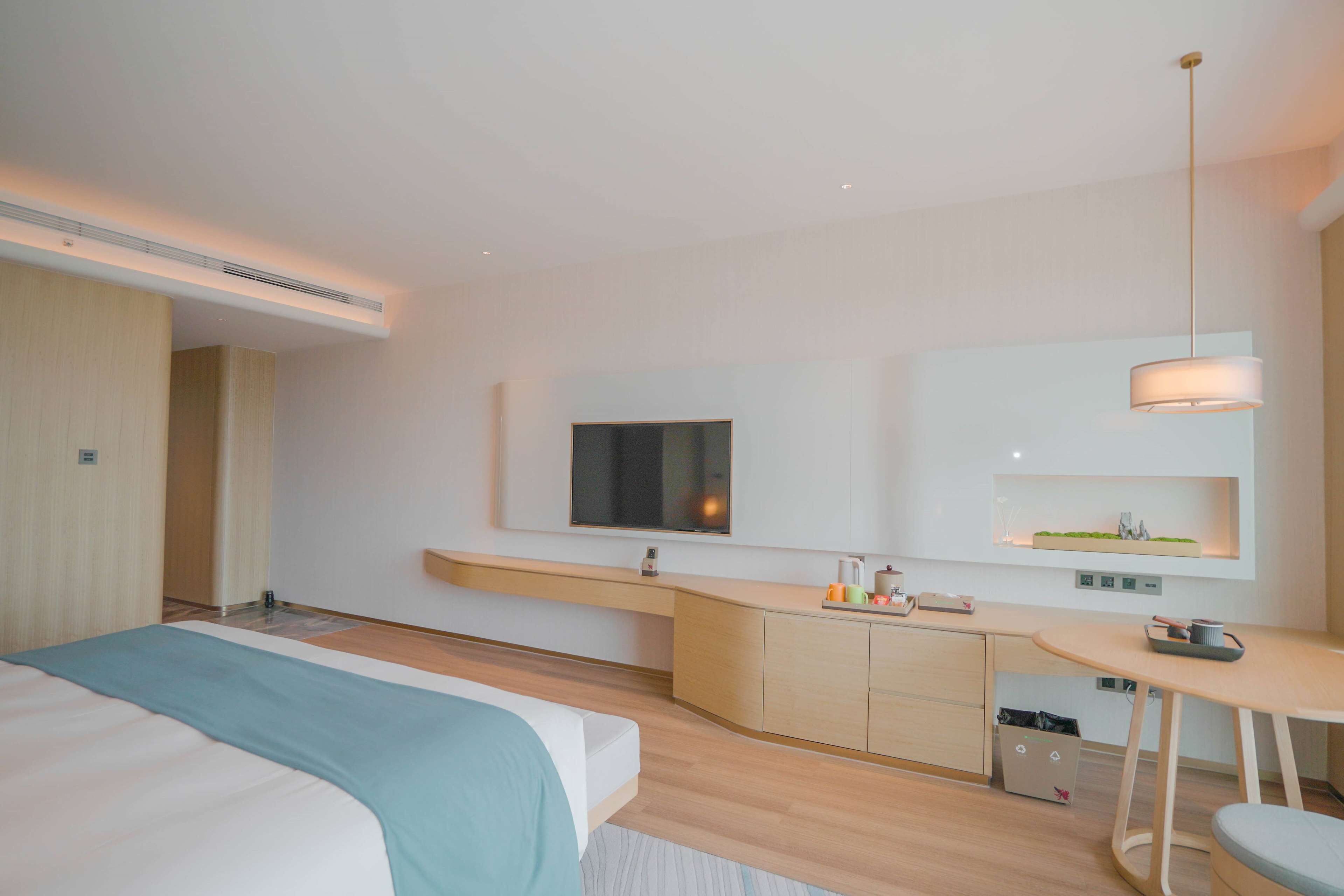 Una habitación de hotel moderna con una cama grande, una kitchenette pequeña, un televisor montado en la pared y un lámpara colgante.