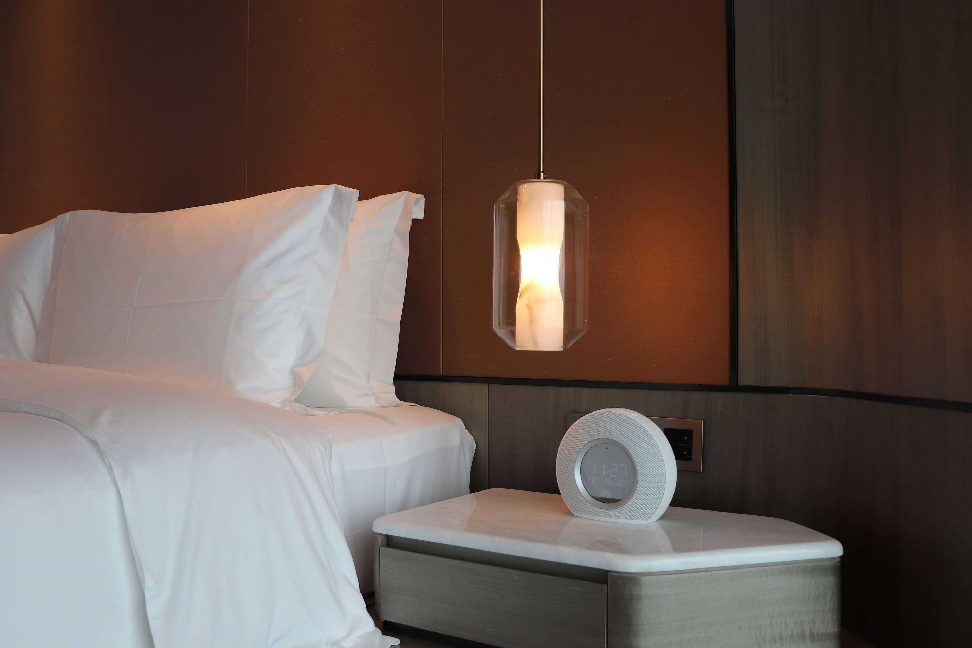 A modern hotel room with a neatly made bed, a bedside table with a digital clock, and a hanging pendant light.