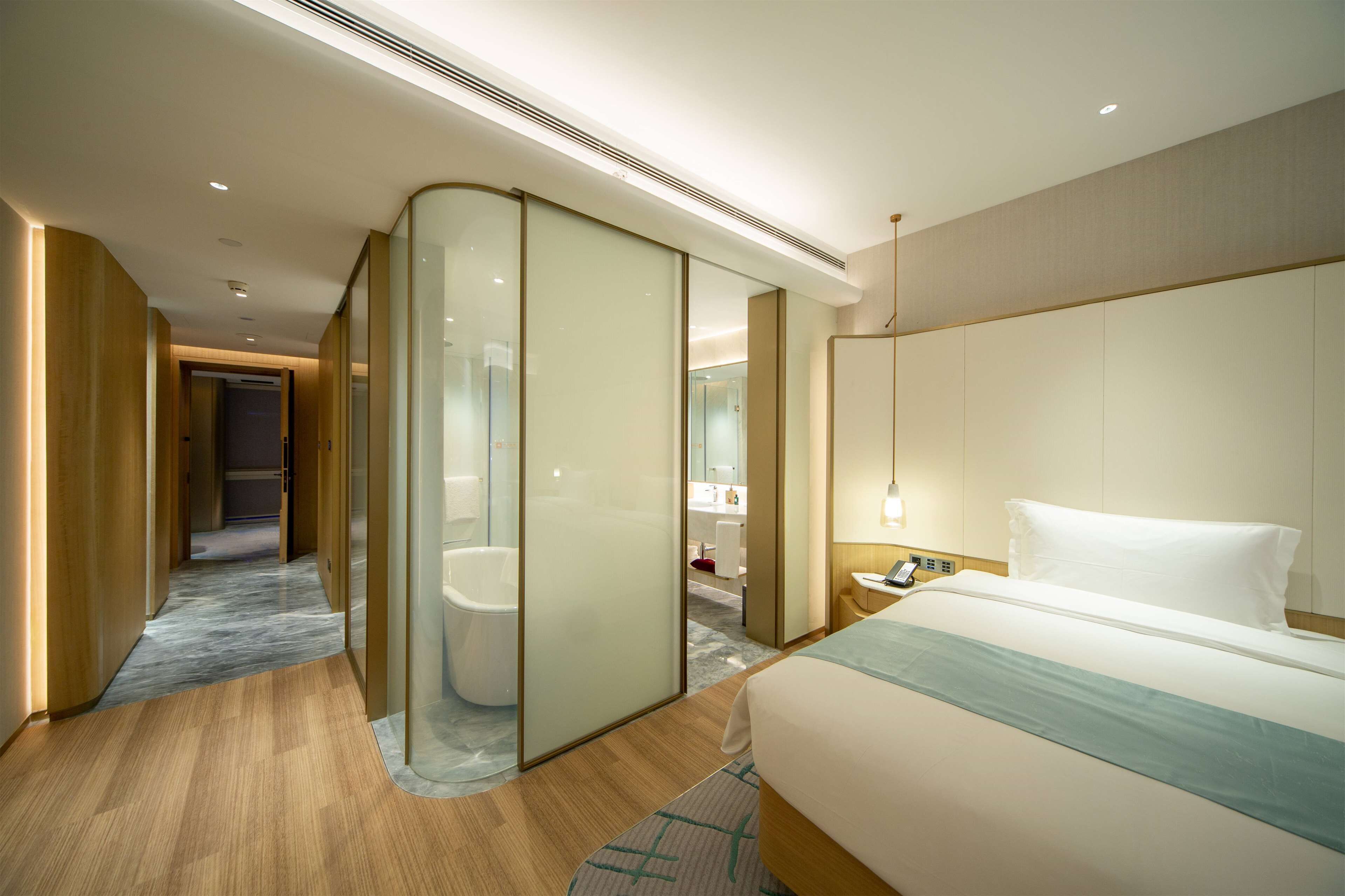 Una habitación de hotel moderna con una cama grande, un separador de vidrio, un baño y un pasillo que conduce a otra habitación.