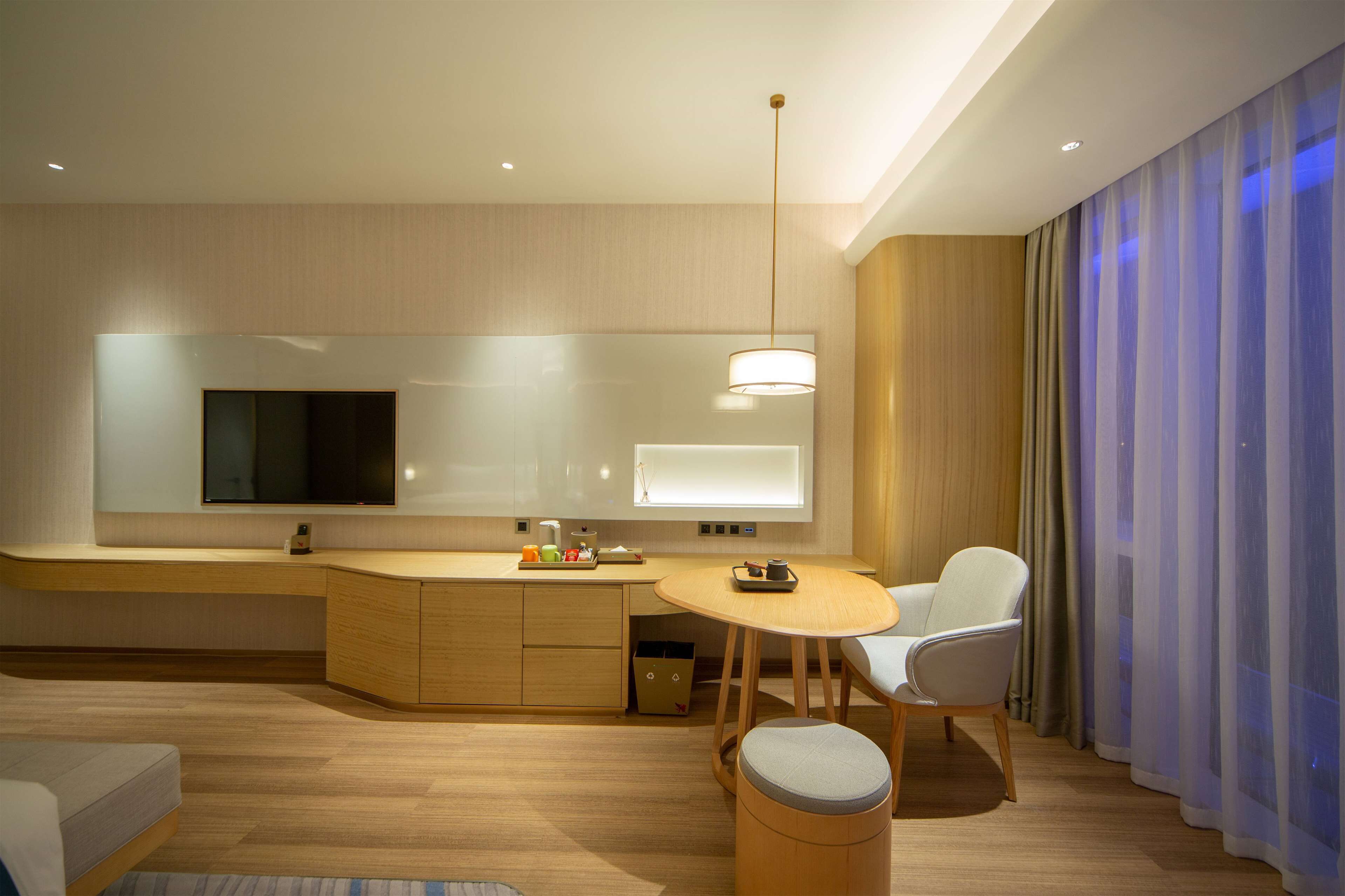 A modern hotel room with a wooden floor, a round dining table, a sofa, a TV mounted on the wall, and a window with sheer curtains.