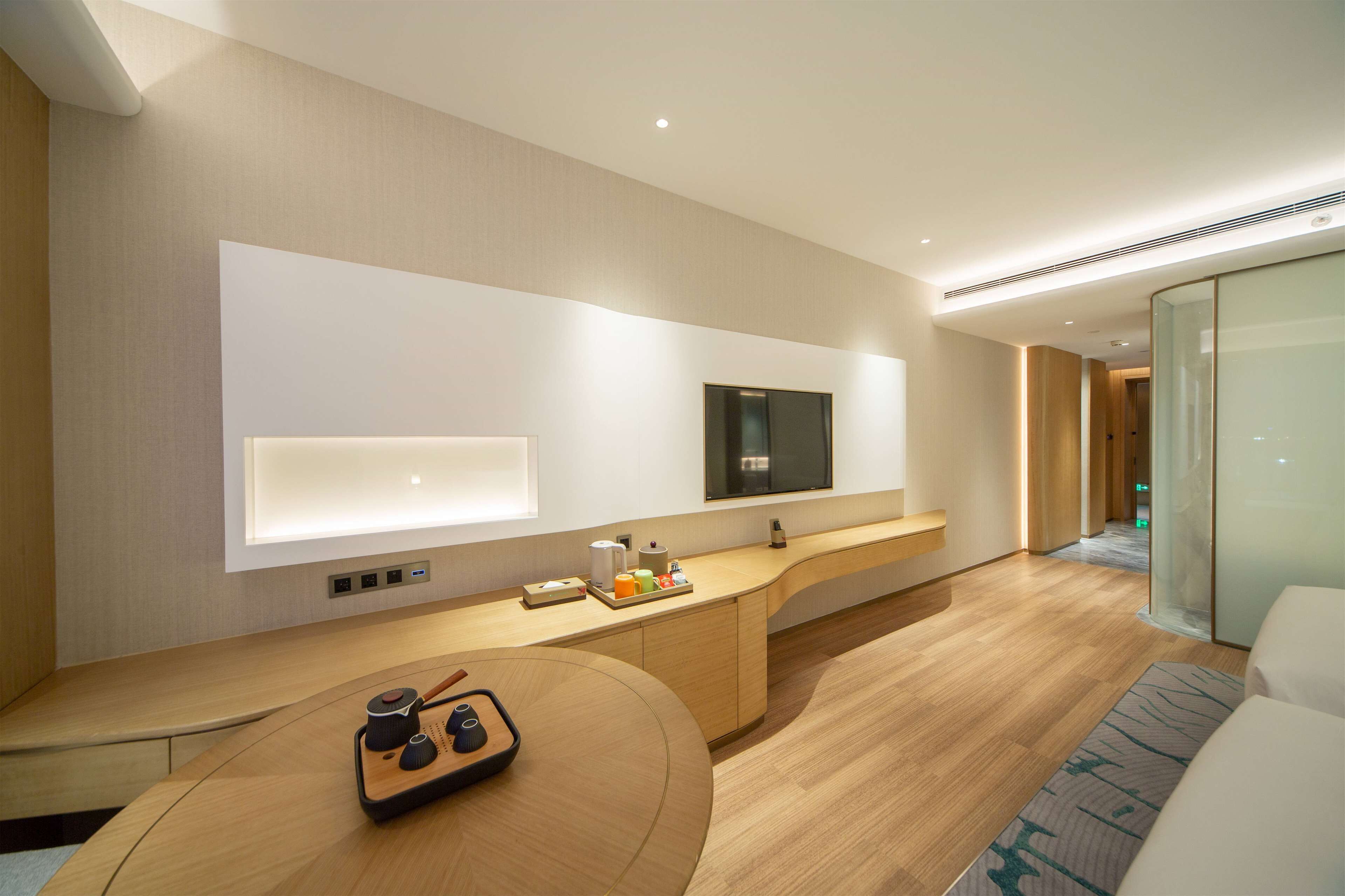Habitació d'hotel moderna amb terra de fusta, una taula consola corba amb joc de te, un televisió de pantalla plana penjada a la paret i una separació de vidre que divideix l'habitació del passadís.