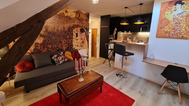 Un loft moderno con techo inclinado, una zona de estar acogedora con un sofá oscuro, una mesa de centro de madera y una alfombra roja. Se ve una cocina con electrodomésticos modernos y luces colgantes al fondo.