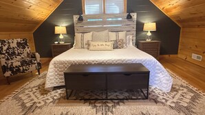 Un dormitorio acogedor con techo inclinado, una cama grande con cabecera, dos lámparas de noche, un banco y una alfombra estampada.