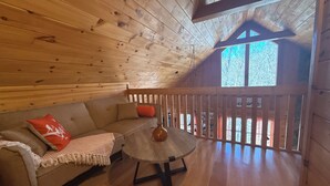 Interior acogedor de una cabaña con techo de madera, un sofá con un cojín rojo, una mesita y un balcón con vistas a árboles.