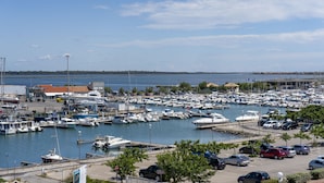 Un puerto deportivo con numerosas embarcaciones amarradas, un estacionamiento con coches y una vista despejada del agua y la costa lejana.