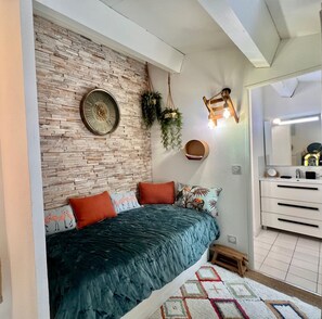 Une chambre avec un mur en briques, un lit recouvert d’une courtepointe bleue, deux oreillers, une plante suspendue, un luminaire fixé au mur et un petit tabouret en bois.