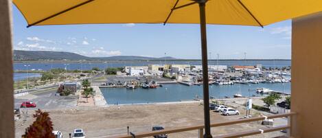 A view from a balcony with a yellow umbrella, overlooking a marina with boats, a parking lot, and a distant hilly landscape.