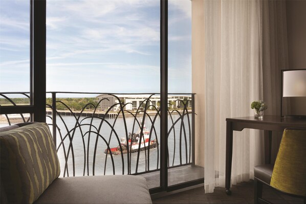 A room with a view of a river, a balcony with a railing, a sofa, a table with a lamp, and a window with curtains.