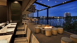 Een terras met tafels en stoelen, een vuurplaats en uitzicht op water en gebouwen.