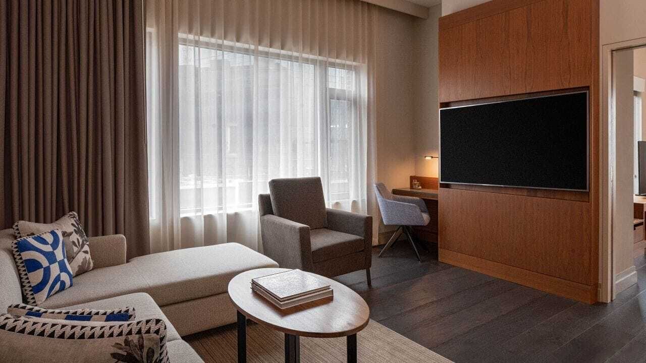 A modern hotel room with a sofa, armchair, and a small table with a book. There is a large flat-screen TV mounted on the wall and a desk with a chair.