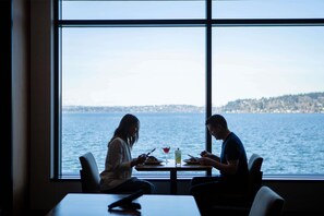A dining area with a view of the water, a table, chairs, and a person using a smartphone. - 2 Connecting Suites at a 4⭐️ Hotel (Renton)