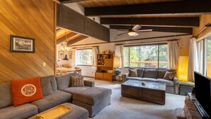 Un salón amplio con un sofá grande, una mesa de centro y una televisión. Se ve una estantería y un comedor al fondo.