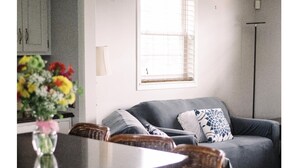 Un espace de vie compact comprenant un coin cuisine, un canapé gris agrémenté de coussins à motifs et un coin repas avec des chaises en osier.