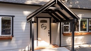 A house with a white exterior, a black roof, and a welcome sign above the door. - Hot Tub + Deck Dining • Mins to State Park Trails + Homewood Ski | Sweet Pine (Tahoma)