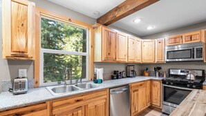 A modern kitchen with wooden cabinets, stainless steel appliances, and a large window providing natural light. - Hot Tub + Deck Dining • Mins to State Park Trails + Homewood Ski | Sweet Pine (Tahoma)