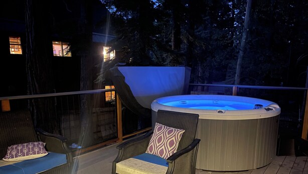 A hot tub on a wooden deck with wicker chairs and cushions, surrounded by trees. - Hot Tub + Deck Dining • Mins to State Park Trails + Homewood Ski | Sweet Pine (Tahoma)