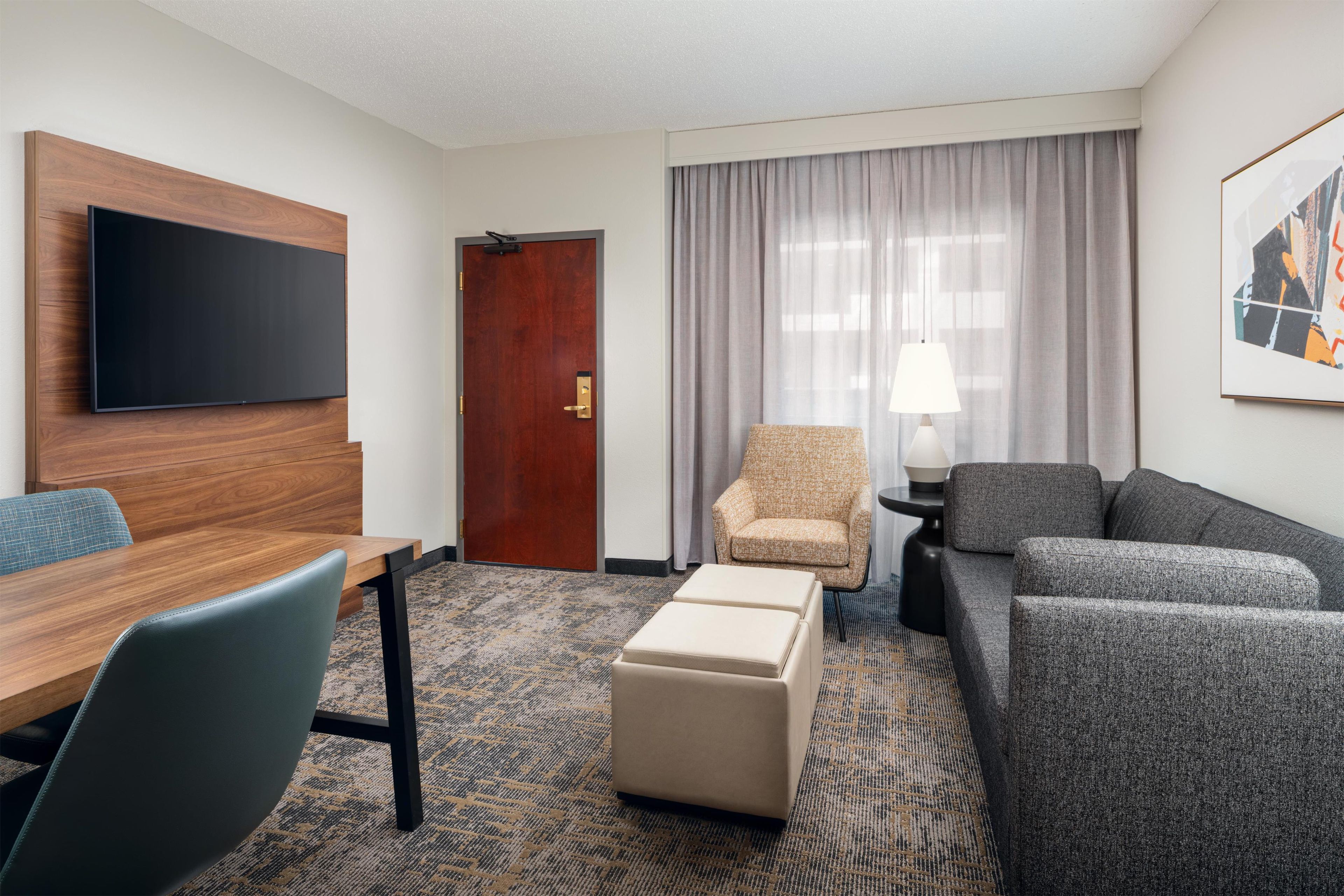 A hotel room with a flat-screen TV mounted on the wall, a wooden desk, a blue chair, a grey sofa, a beige ottoman, a patterned carpet, a door with a brass handle, and a window with sheer curtains.