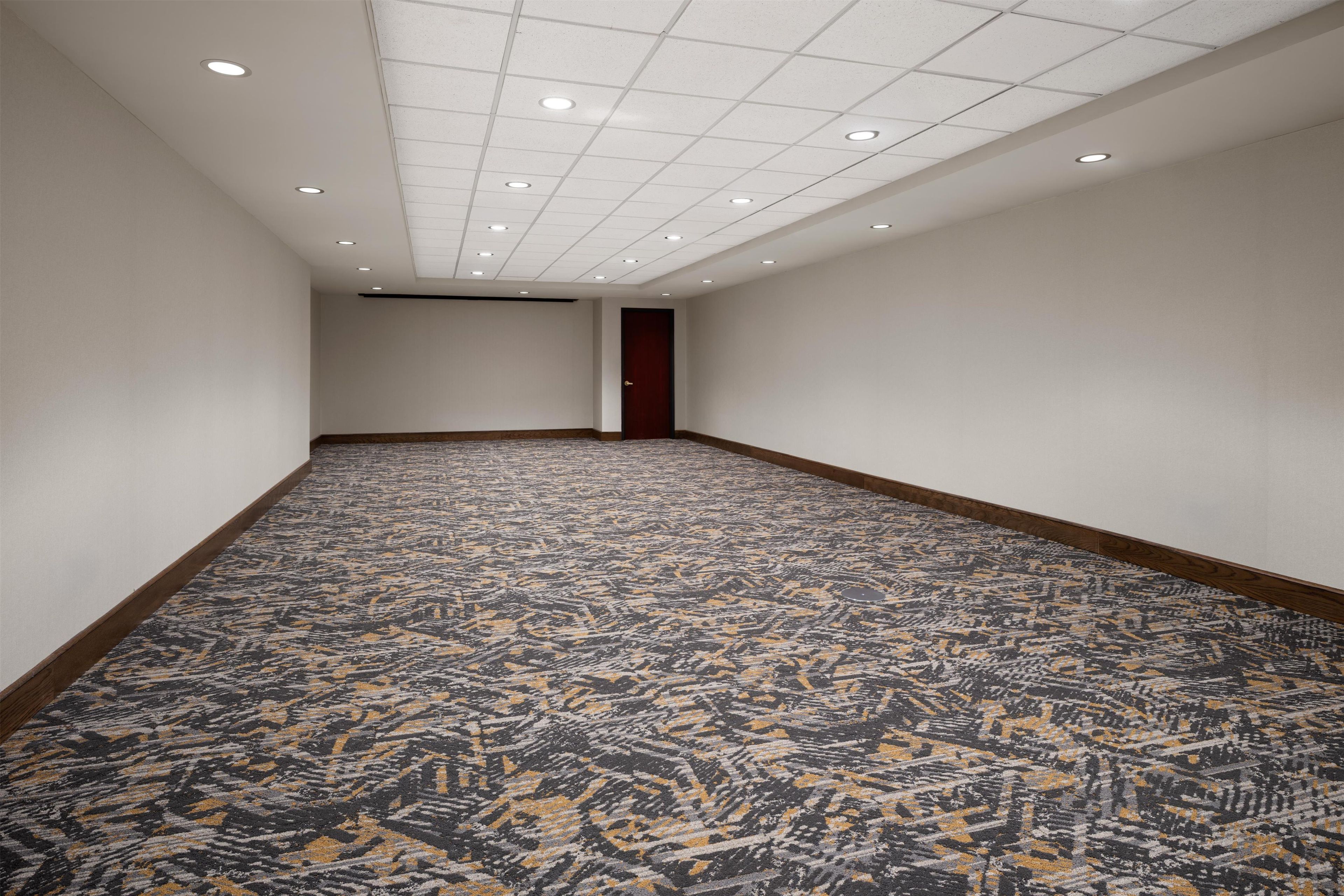 A long, narrow hotel room with a patterned carpet, white walls, and a wooden door.