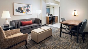 A hotel room with a gray sofa, a wooden dining table, a patterned rug, and a kitchenette in the background. - 2 Connecting Suites at a  Hotel (Lincoln)