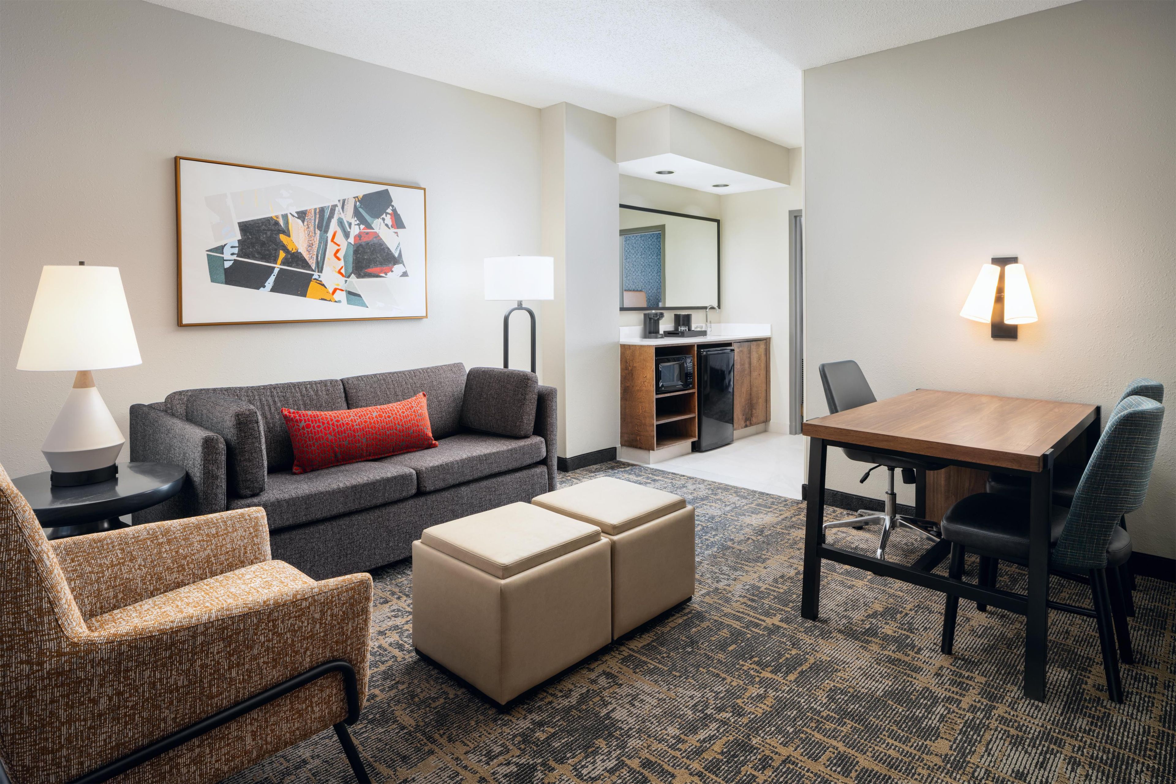 A hotel room with a gray sofa, a wooden coffee table, a patterned armchair, a small ottoman, a dining table with chairs, a kitchenette, and a large framed abstract painting.