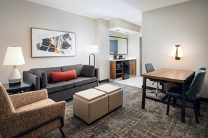 Habitación de hotel con sofá gris, una mesa de centro de madera, un sillón estampado, un pequeño otoman, una mesa de comedor con sillas, una kitchenette y un cuadro abstracto grande enmarcado.