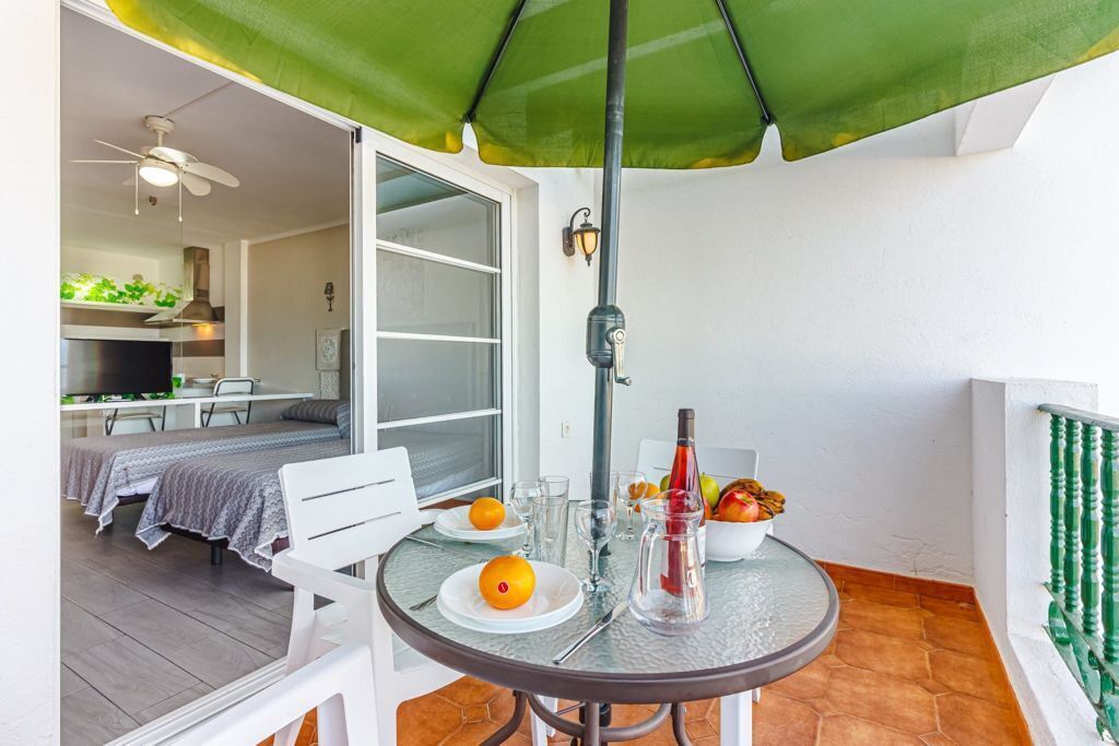 A balcony with a table set for two, a green umbrella, and a kitchen in the background.
