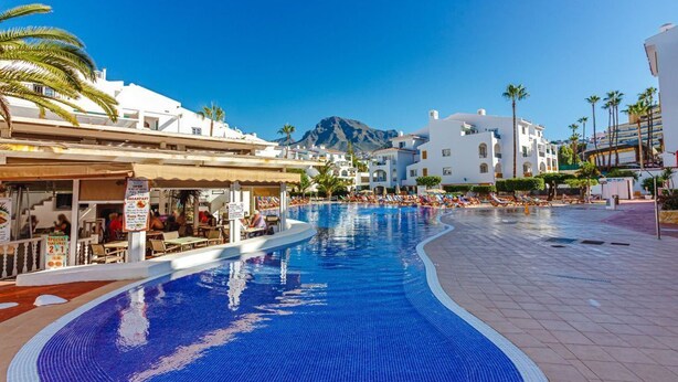 A swimming pool with lounge chairs, a restaurant, and buildings with white facades and palm trees. - Spectacular apartment with sunset ocean views in Fanabe Beach with free WIFI (Costa Adeje)
