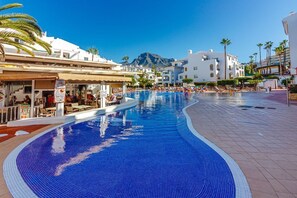 Ein Swimmingpool mit Liegestühlen, ein Restaurant und Gebäude mit weißen Fassaden und Palmen.