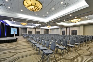 Un salón de conferencias con filas de sillas, un escenario con una pantalla y varias luces en el techo.