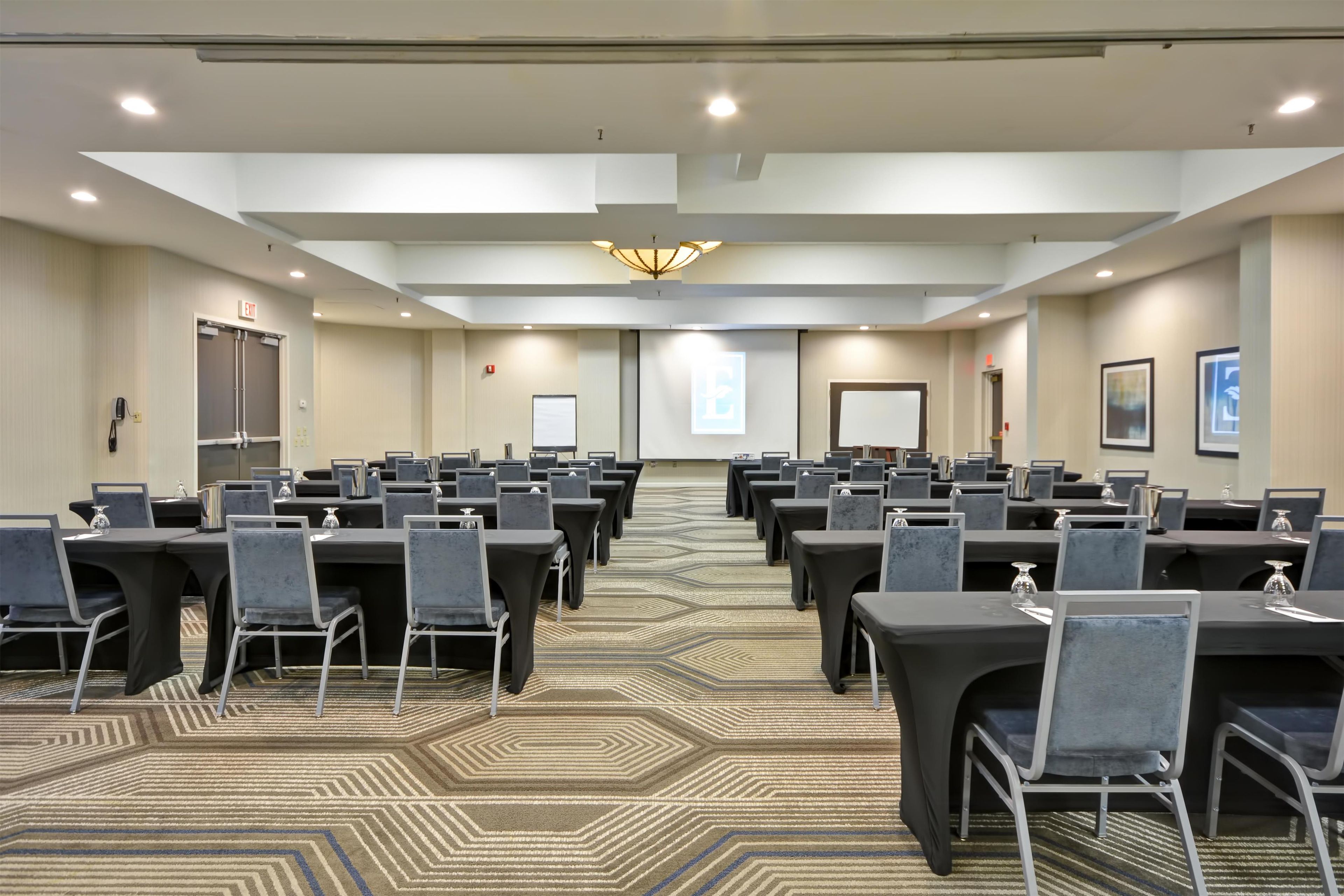 A conference room with rows of tables and chairs, a patterned carpet, and a projector screen.