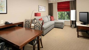 Une chambre d’hôtel avec un bureau en bois, un canapé gris avec des coussins rouges, une télévision et une fenêtre avec des rideaux rouges.
