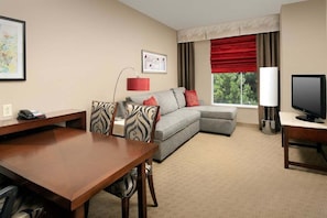 Quarto de hotel com uma secretária de madeira, um sofá cinzento com almofadas vermelhas, uma televisão e uma janela com cortinas vermelhas.