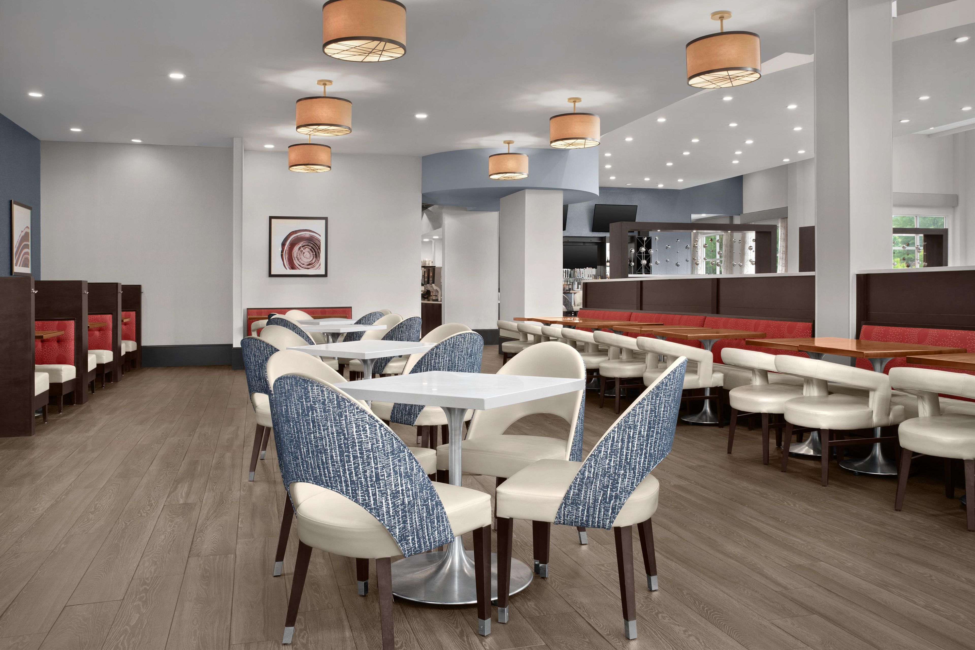 A modern restaurant interior with multiple dining tables and booths, overhead pendant lighting, and a visible kitchen area.