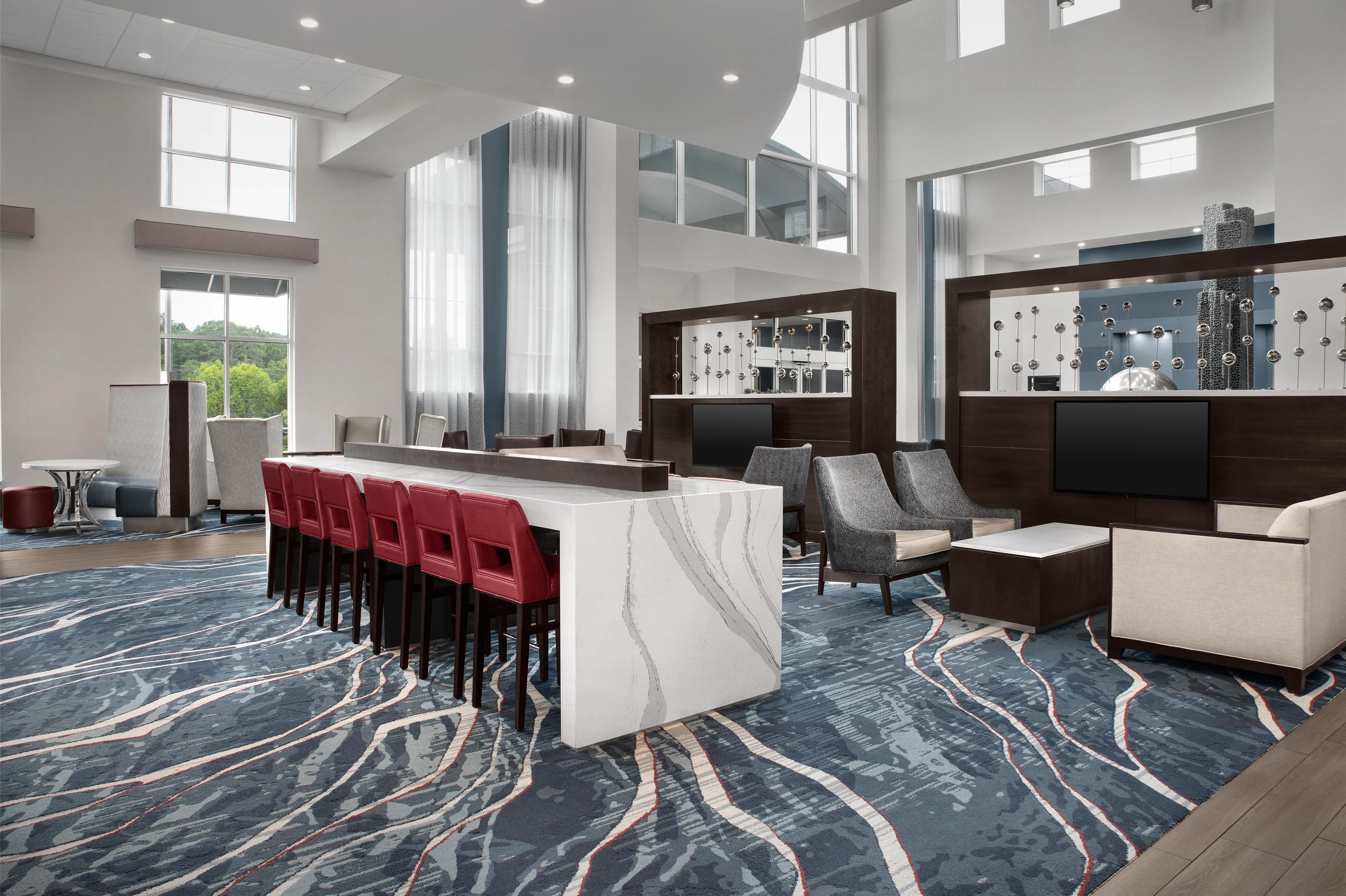 Un moderno vestíbulo de hotel con mostrador de recepción de mármol, taburetes de bar rojos, una zona de estar con alfombra estampada y un gran ventanal con vistas al exterior.