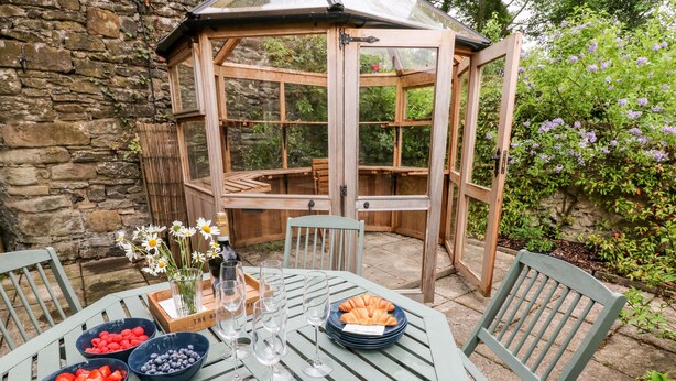 A wooden table set with fruit bowls, wine glasses, and a bottle of champagne, surrounded by green chairs in a garden with a stone wall and a glass greenhouse structure. - Eastview (Matlock)