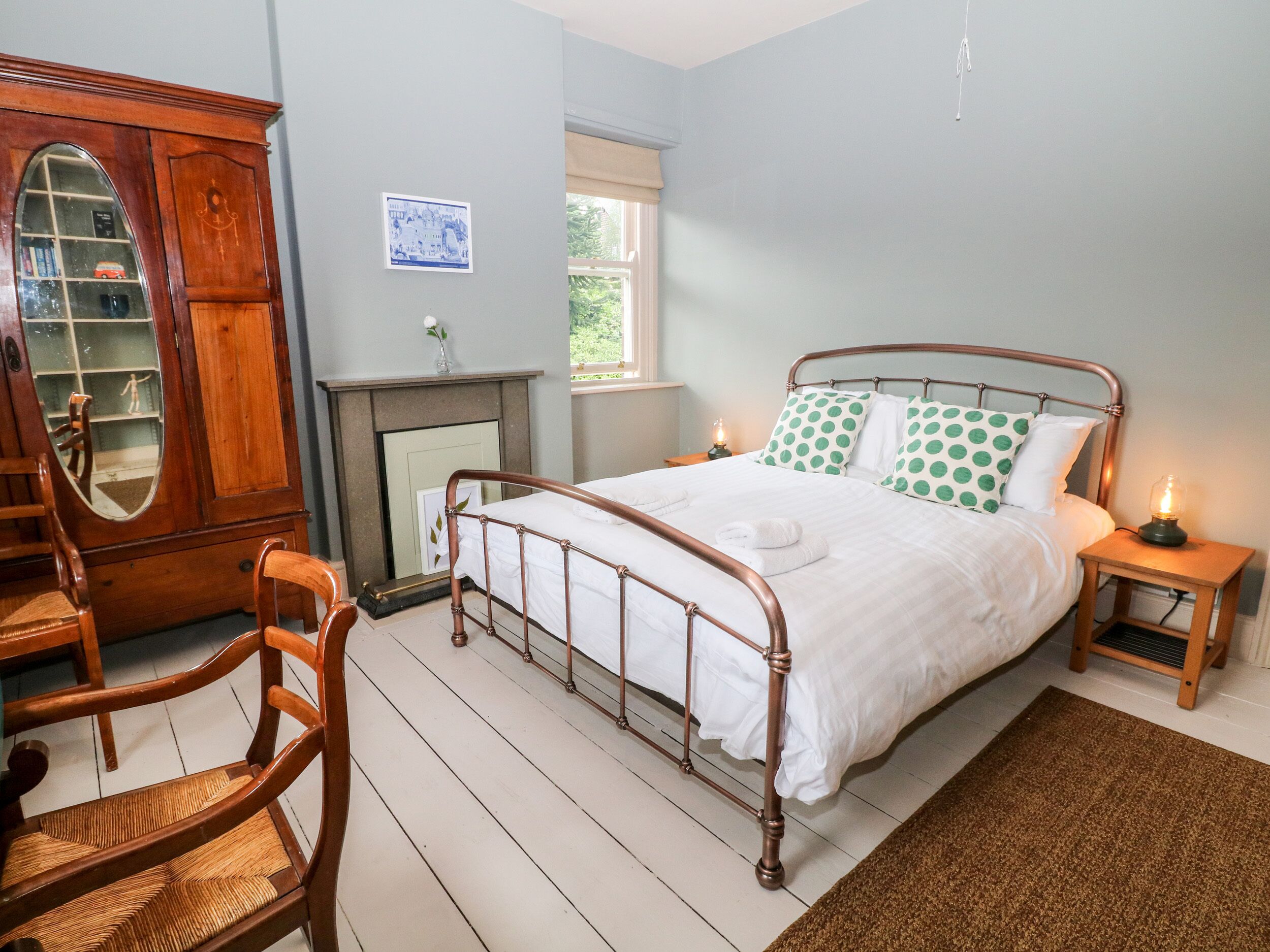 A bedroom with a metal-framed bed, a wooden chair, a wooden cabinet with a mirror, and a fireplace.