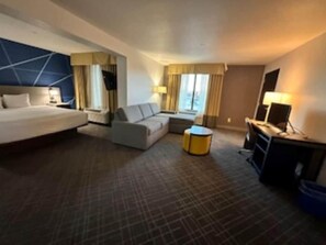 A hotel room with a bed, sofa, armchair, desk, and a small round table. - 2 Connecting Suites at a  Hotel (Frisco)