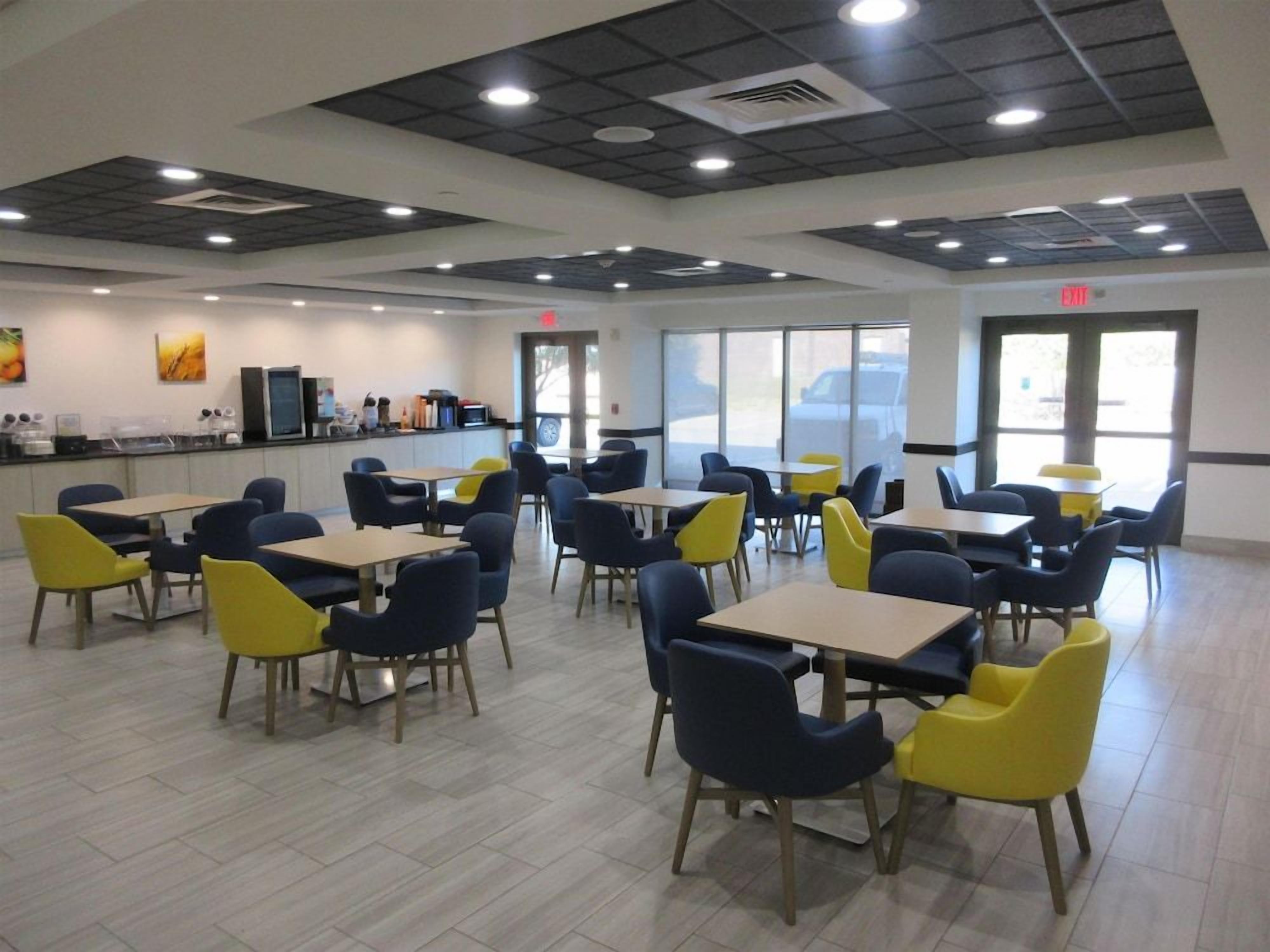 A cafeteria with tables and chairs, a counter with food service equipment, and large windows.