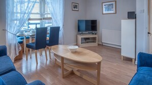 Una sala de estar con una mesa redonda de madera, sillas azules, un televisor sobre un mueble bajo y un radiador.