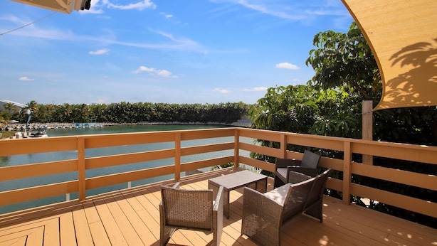 Une terrasse en bois, meublée de meubles en osier, donnant sur une étendue d’eau et entourée de verdure.