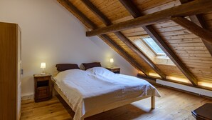 Ein Schlafzimmer mit schräg versiegelter Decke, Holzbalken, einem Bett mit weißer Bettwäsche, zwei Nachttischen mit Lampen sowie einem Oberlicht.