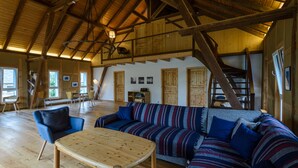 Ein großzügiger Wohnbereich mit hoher Holzbalkendecke, einem großen gestreiften Sofa, einem Holztisch und einer Treppe, die in eine obere Ebene führt.