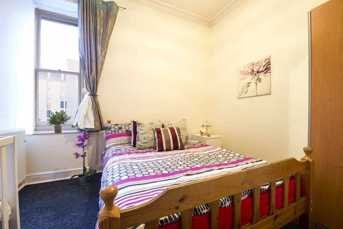 A bedroom with a wooden bed, a nightstand, a window with curtains, a potted plant, and a wall-mounted flower painting.