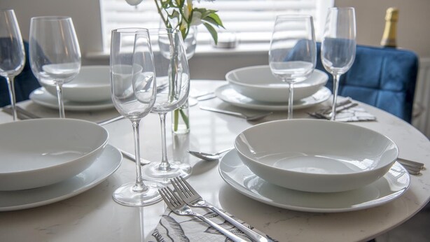 Une table à manger dressée avec de l’argenterie, des verres à vin et des assiettes blanches, et un vase de fleurs placé au centre.