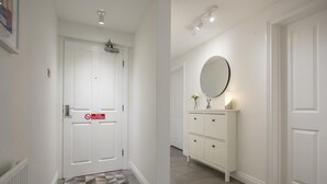 Un couloir avec une porte blanche, un panneau «interdiction de fumer», une commode blanche avec des tiroirs, un miroir rond et un petit vase avec des fleurs.