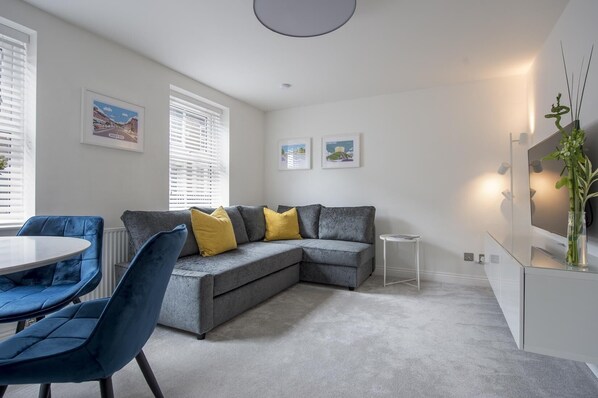 A modern living room with a grey sectional sofa, blue chairs, a small round table, and a white cabinet with a mirror. - Penleys Apartment (York)