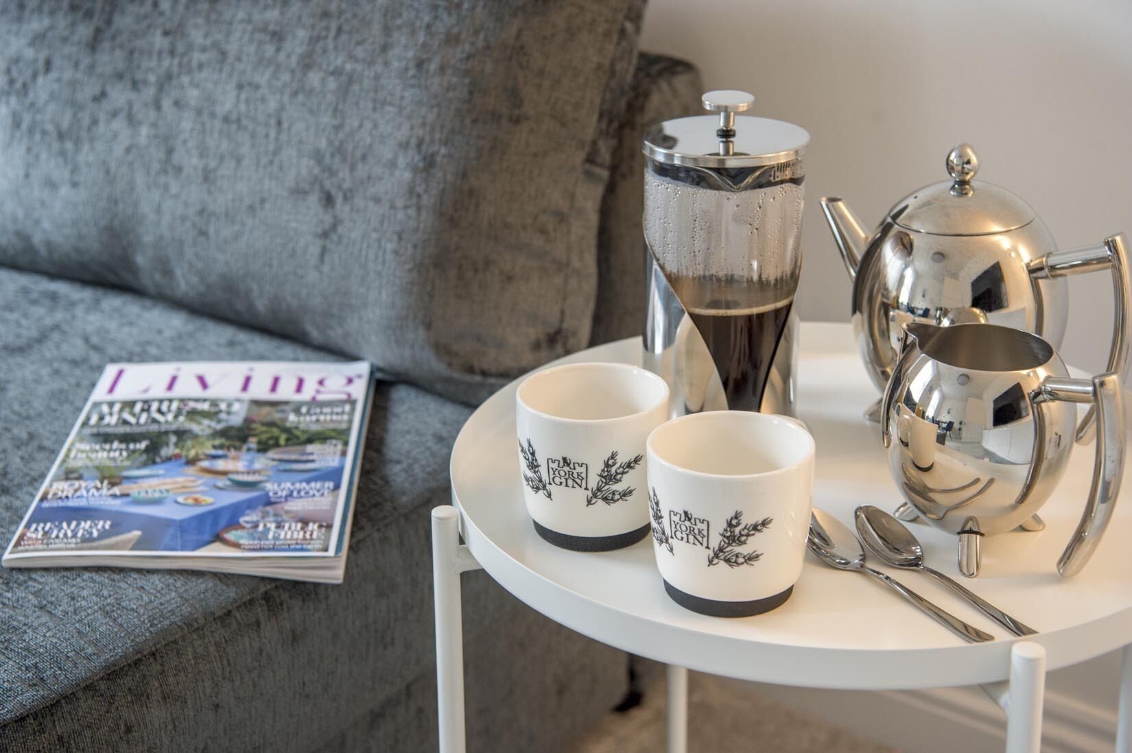 Un tavolo con una rivista, due tazze bianche con decorazioni nere, un teiera d'argento e una caffettiera d'argento.