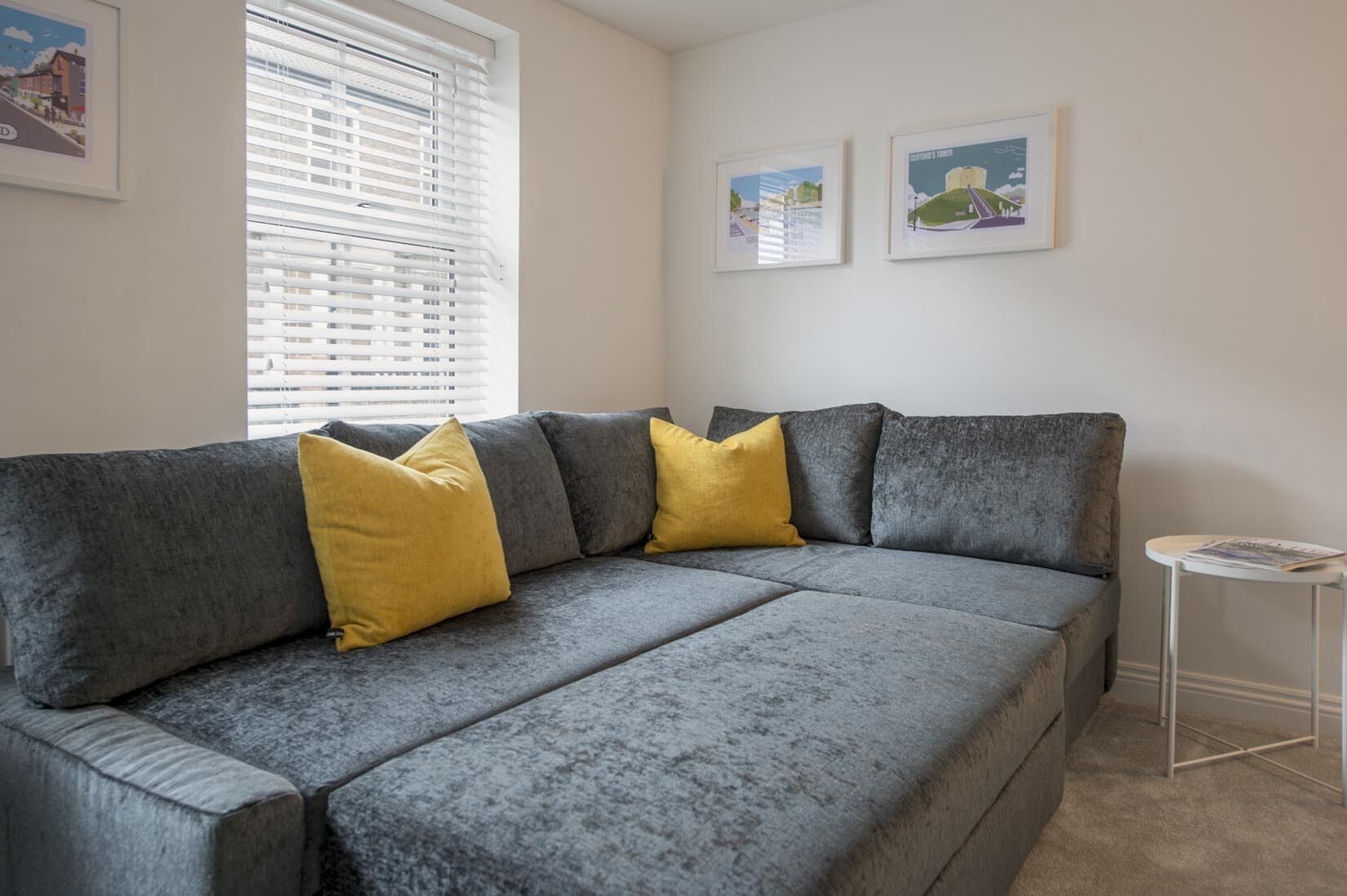 A grey sofa with yellow cushions, a small white side table, and two framed pictures on the wall.