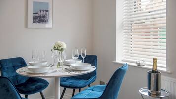 Een eetruimte met een ronde tafel gedekt voor vier personen, blauwe fluweelstoelen, witte borden, wijnglazen en een fles champagne.